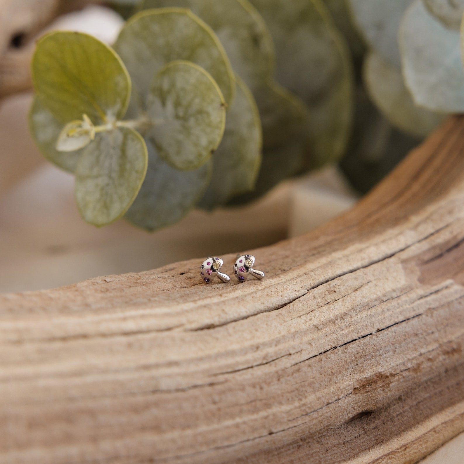 Delicate silver Mushroom Studs with sparkling CZ diamonds rest on a textured wood surface, green leaves in the background—a charming piece of nature-inspired jewelry.