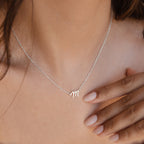 A woman wears the Grace Dainty Initial Necklace, a delicate silver piece featuring an "m" pendant, her hand gently touching her chest.