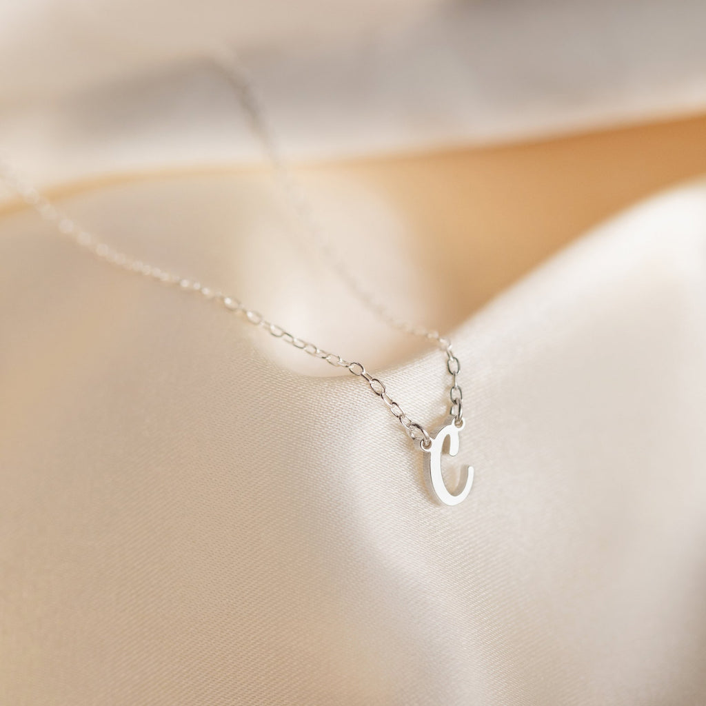 The Grace Dainty Initial Necklace features a silver, small lowercase letter "c" pendant, elegantly displayed on soft, light-colored fabric.
