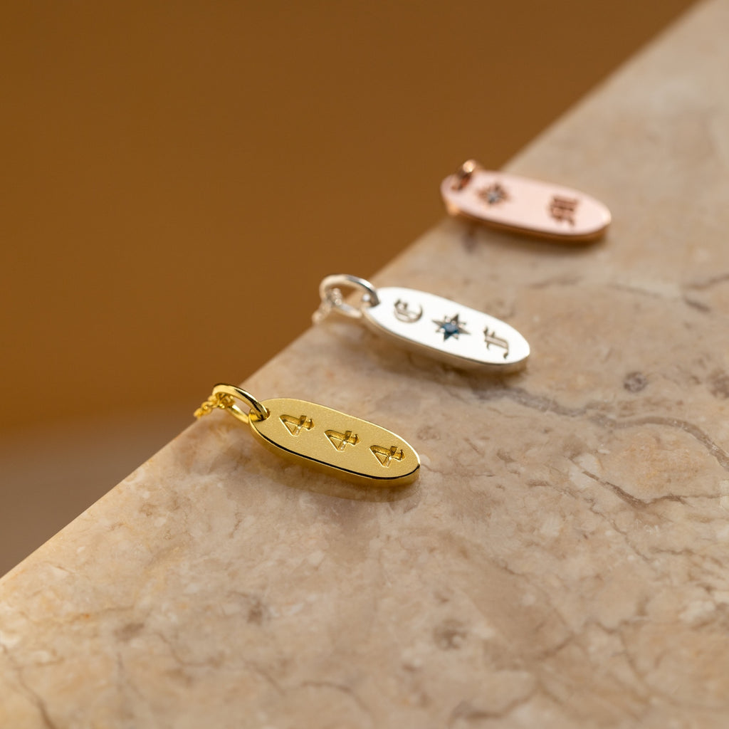 The Old English Oval Tag Necklace features three metal pendants—gold, silver, and rose gold—with engraved initials or symbols, displayed on marble for a personalized touch.