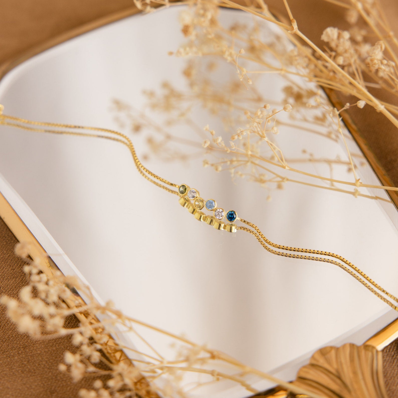 The Cluster Birthstone Bracelet with colorful gemstones is displayed on a mirror, encircled by dried beige flowers.