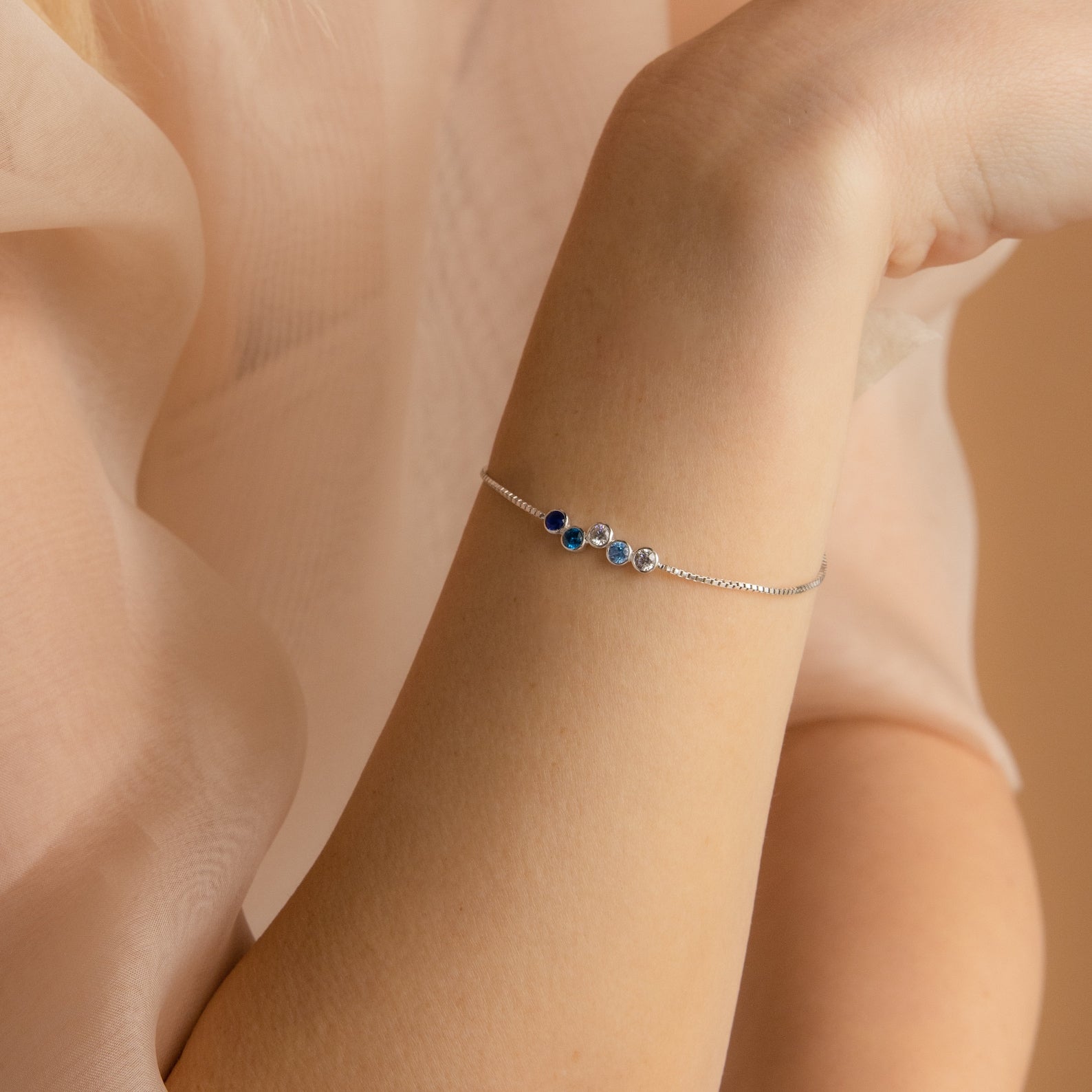 A woman wears the Cluster Birthstone Bracelet—an elegant silver piece with three blue stones—on her wrist, set against a soft beige background.