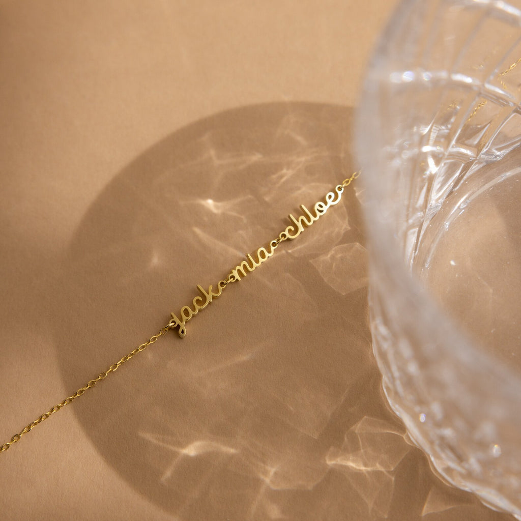 A gold cursive name necklace rests on a beige surface beside a textured glass casting patterns—perfect for minimal jewelry fans or pairing with the Petite Tiny Multiple Name Bracelet for a unique, personalized style.