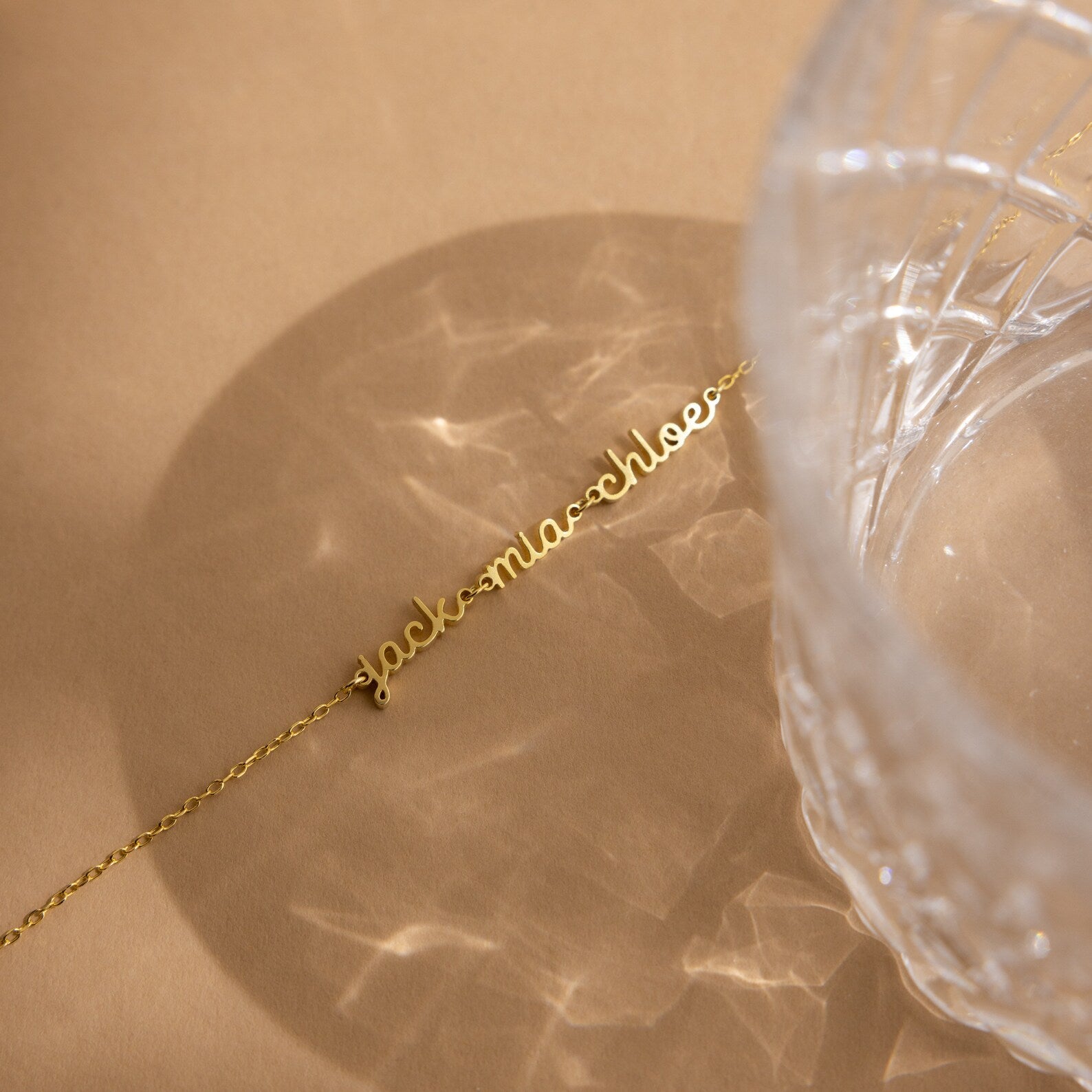 A gold cursive name necklace rests on a beige surface beside a textured glass casting patterns—perfect for minimal jewelry fans or pairing with the Petite Tiny Multiple Name Bracelet for a unique, personalized style.