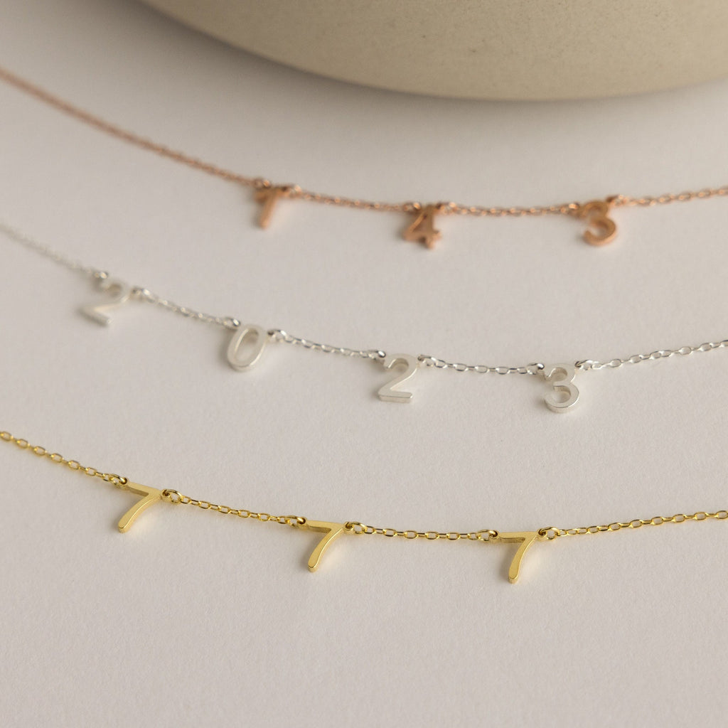 The Alice Spaced Number Necklace collection includes three minimalist necklaces with small number charms: 745 in rose gold, 2023 in silver (Birth Year Necklace), and 777 in gold, displayed on a beige background.