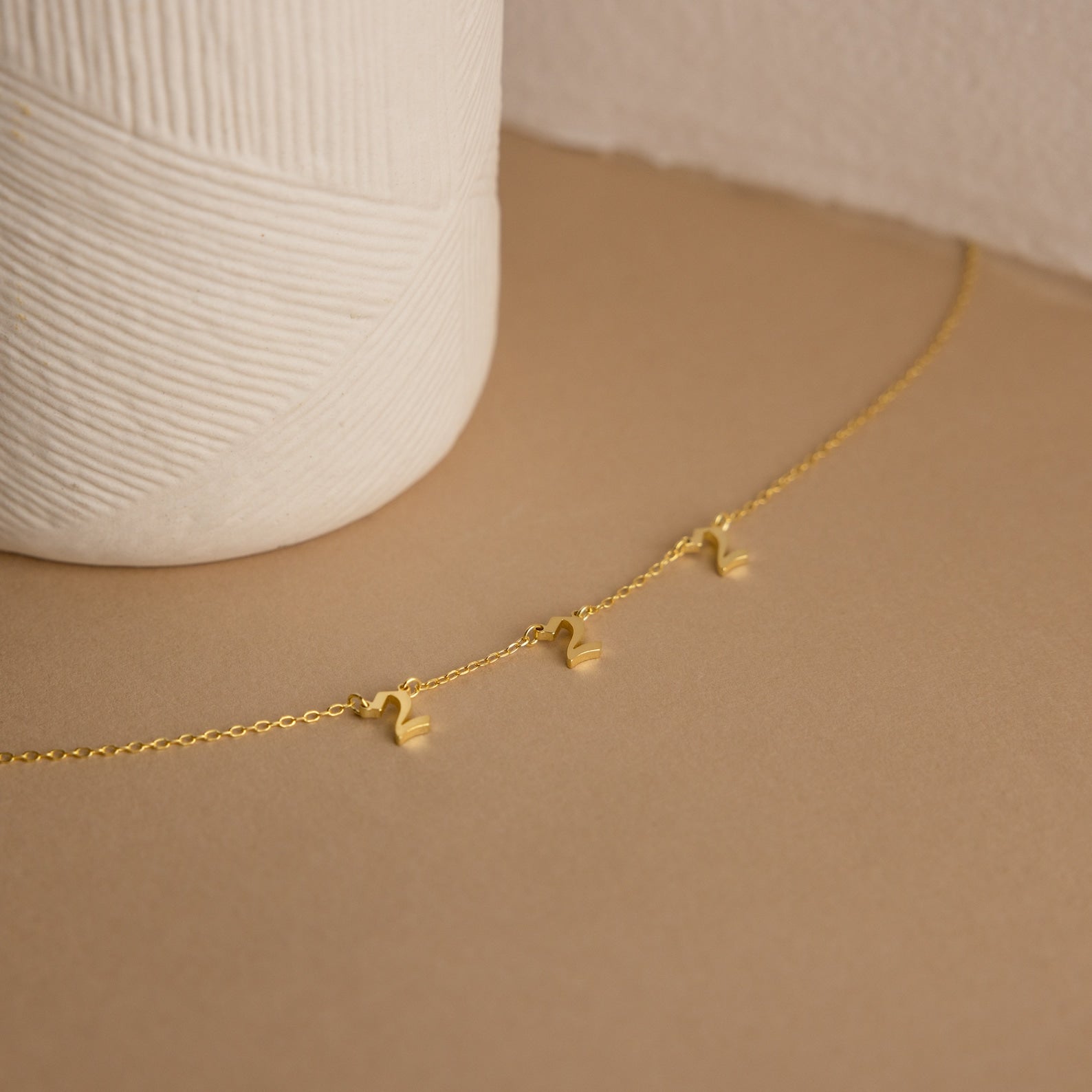 The Old English Spaced Number Necklace, a delicate gold piece featuring spaced number charms, is displayed on a beige surface next to a textured vase.