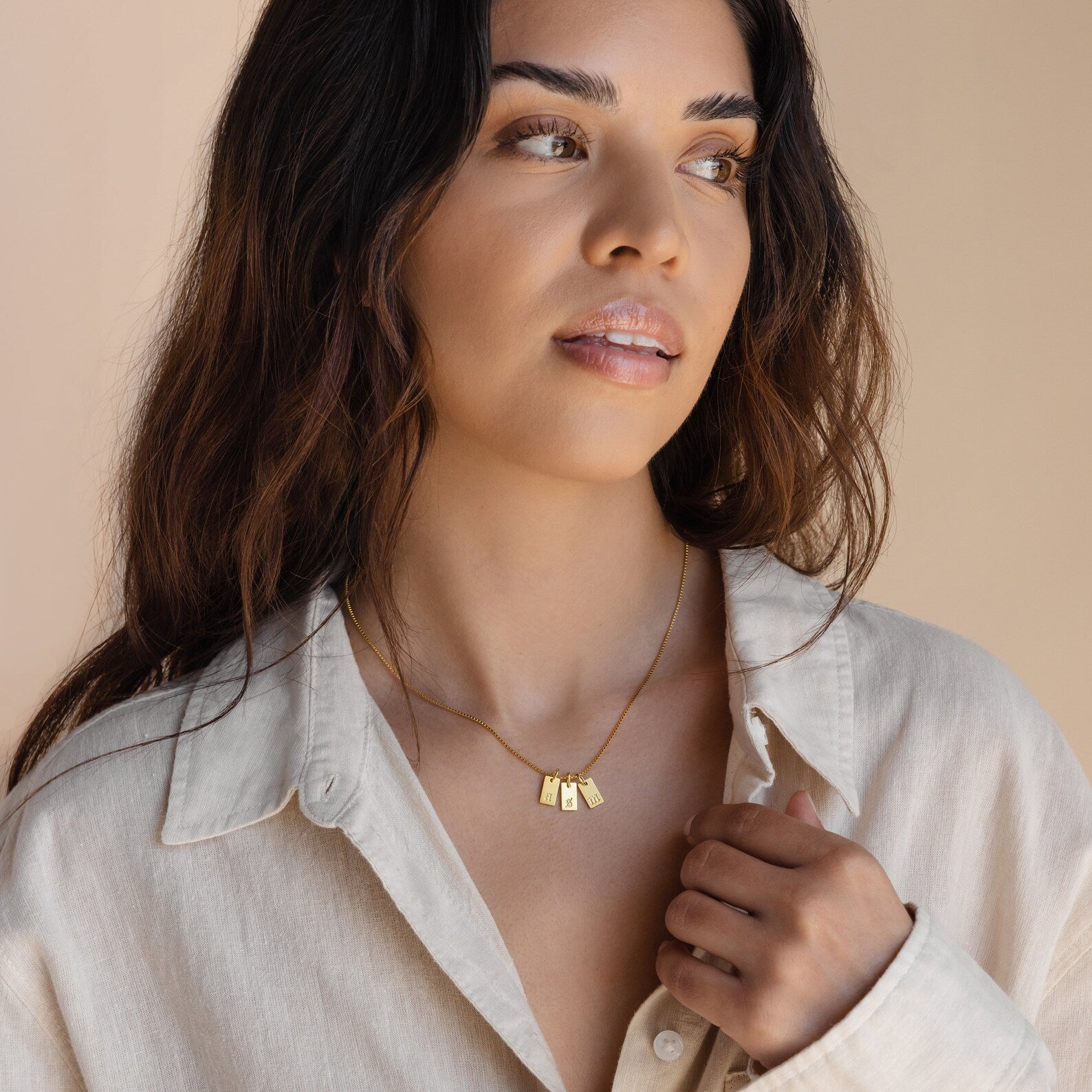 Woman wearing a cream shirt and the Old English Mini Tag Necklace with three rectangular gold pendants, looking off to the side.