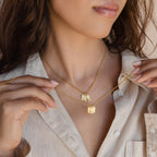 A woman wearing two gold necklaces with rectangular pendants, holding the Carrie Mini Tag Necklace near her beige shirt.