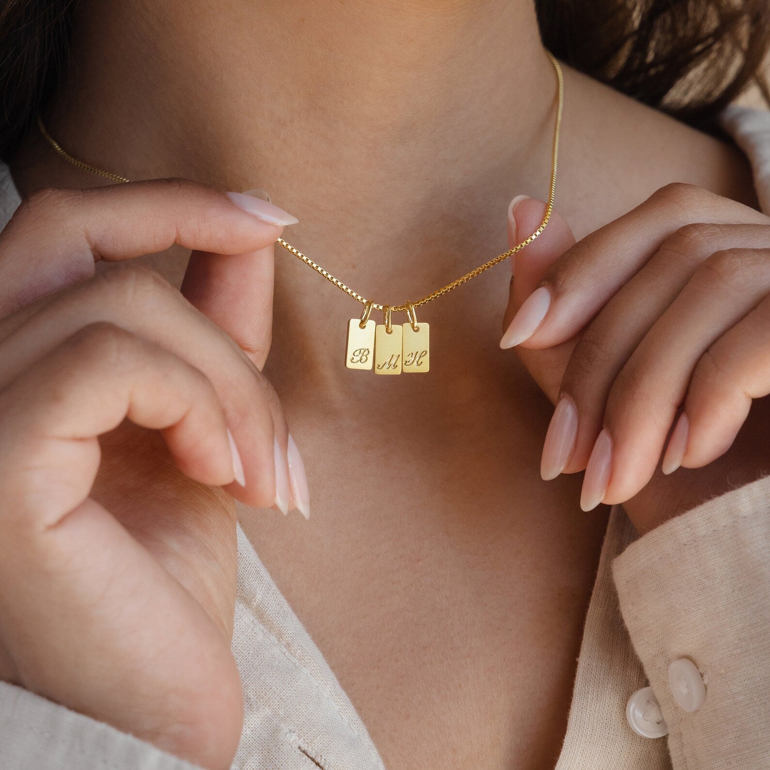 A woman holds the Carrie Mini Tag Necklace—a minimalist, personalized piece featuring three engraved letter pendants: B, A, and L.