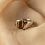 The Tigers Eye Signet Ring in Sterling Silver, featuring a rectangular tiger’s eye stone, is displayed on beige fabric.