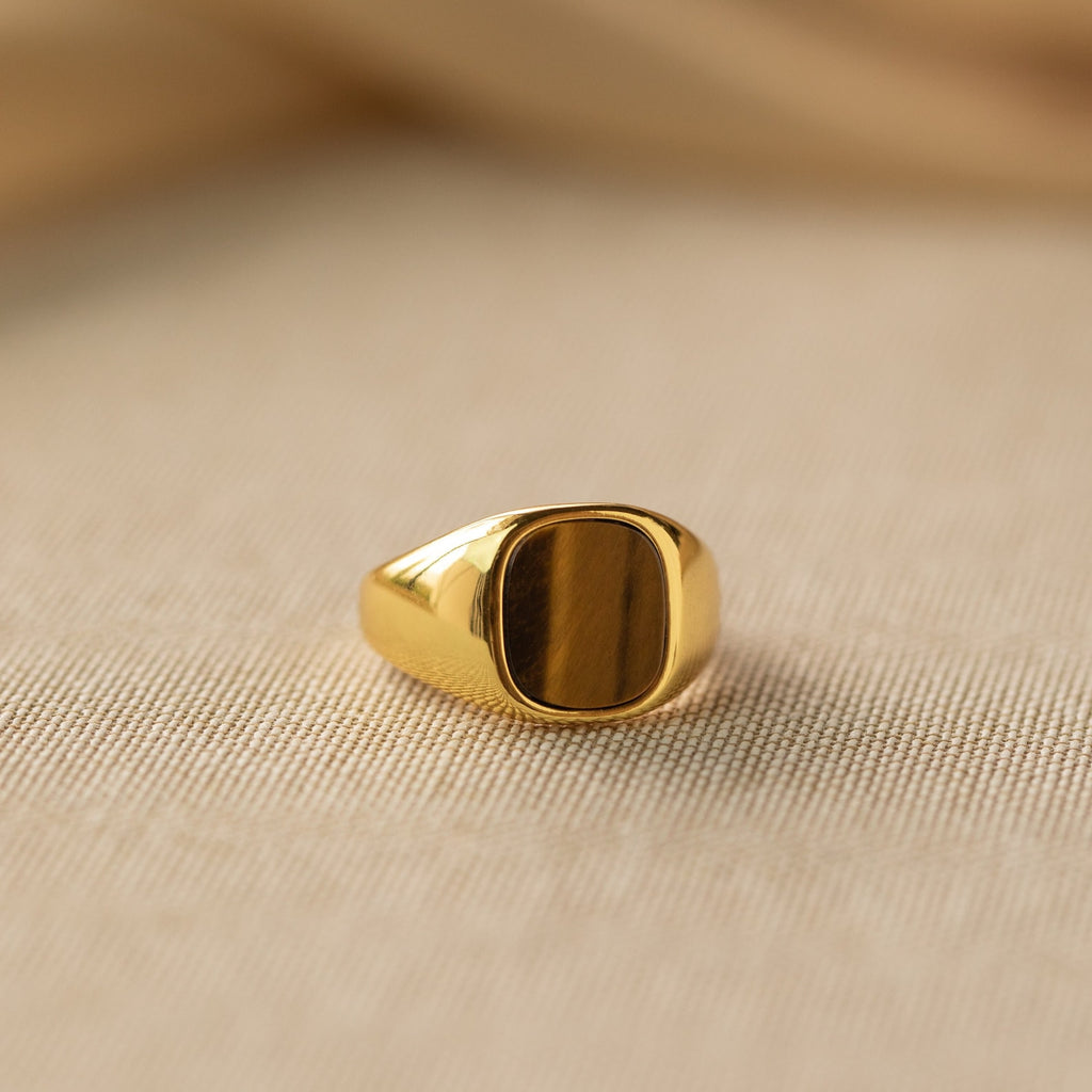 The Tigers Eye Signet Ring in 18K Gold, featuring a rectangular brown stone, is shown on a beige textured surface.