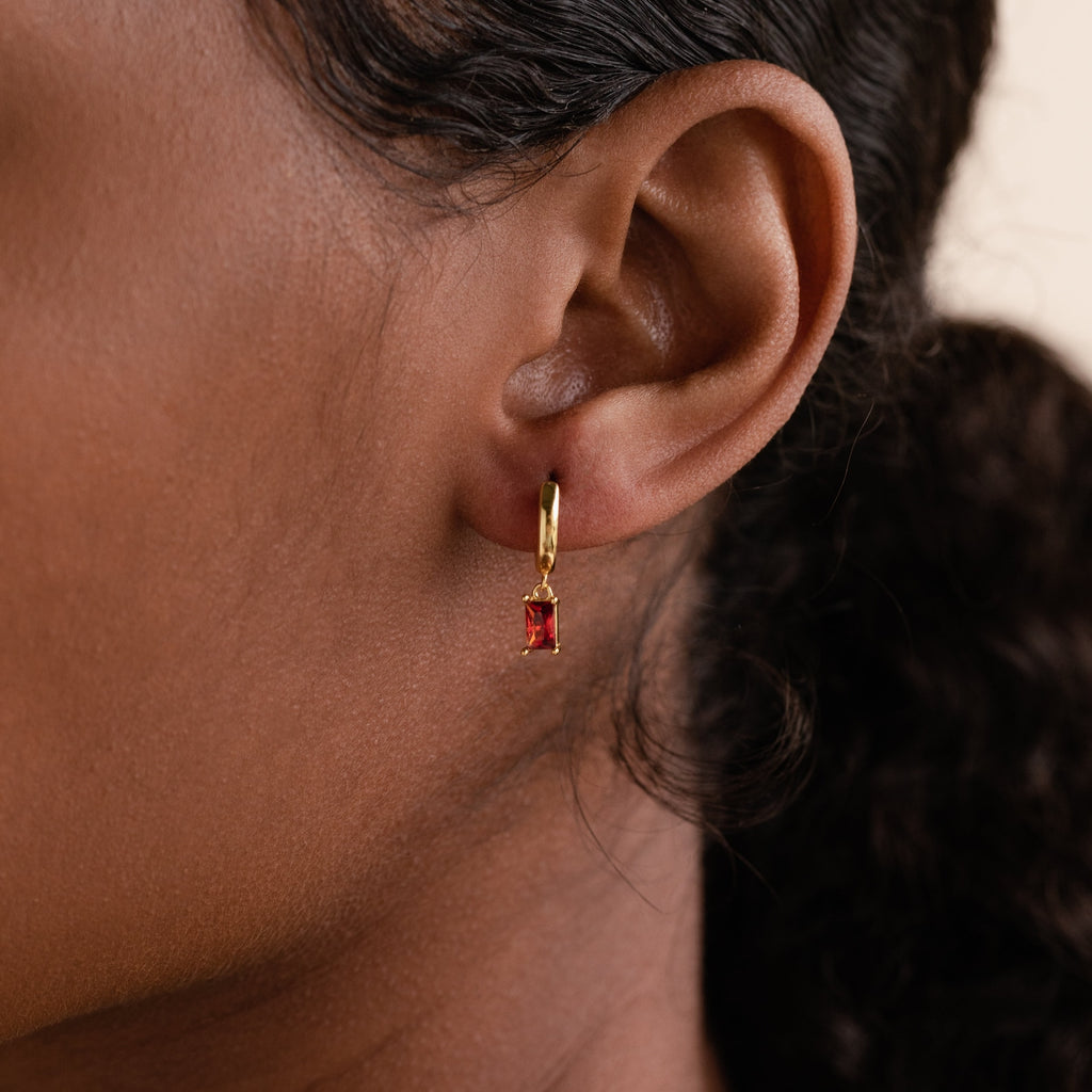 Close-up of a model wearing a gold huggie earring with a deep red baguette-cut garnet charm.