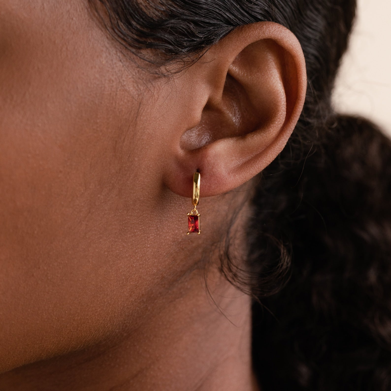 Close-up of a model wearing a gold huggie earring with a deep red baguette-cut garnet charm.