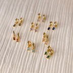 Collection of gold huggie earrings with colorful baguette-cut birthstones, arranged on a pleated ivory fabric background.