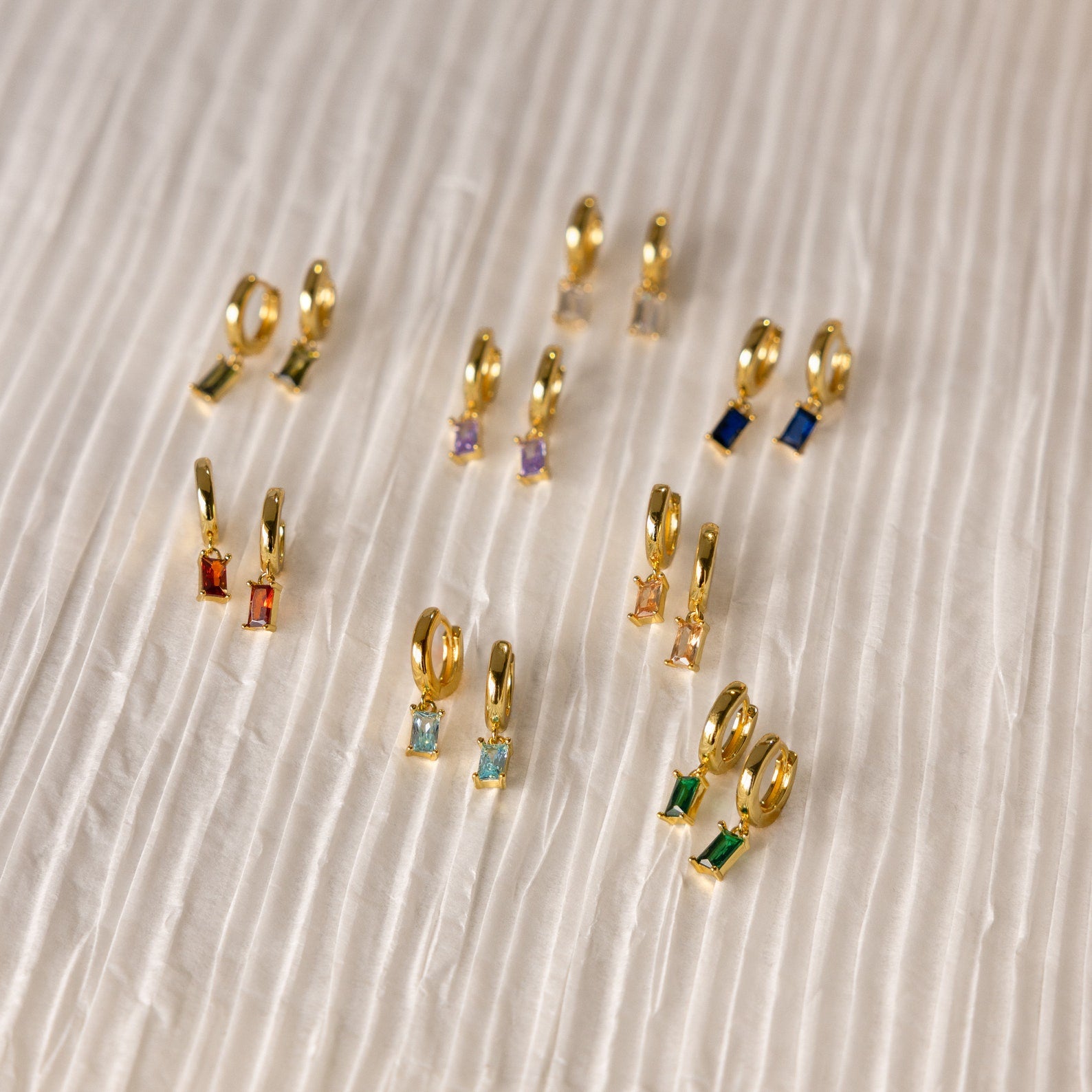 Collection of gold huggie earrings with colorful baguette-cut birthstones, arranged on a pleated ivory fabric background.