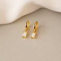 Gold huggie earrings with rectangular opal stones, glowing with iridescent hues, displayed on beige fabric.