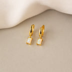 Gold huggie earrings with rectangular opal stones, glowing with iridescent hues, displayed on beige fabric.