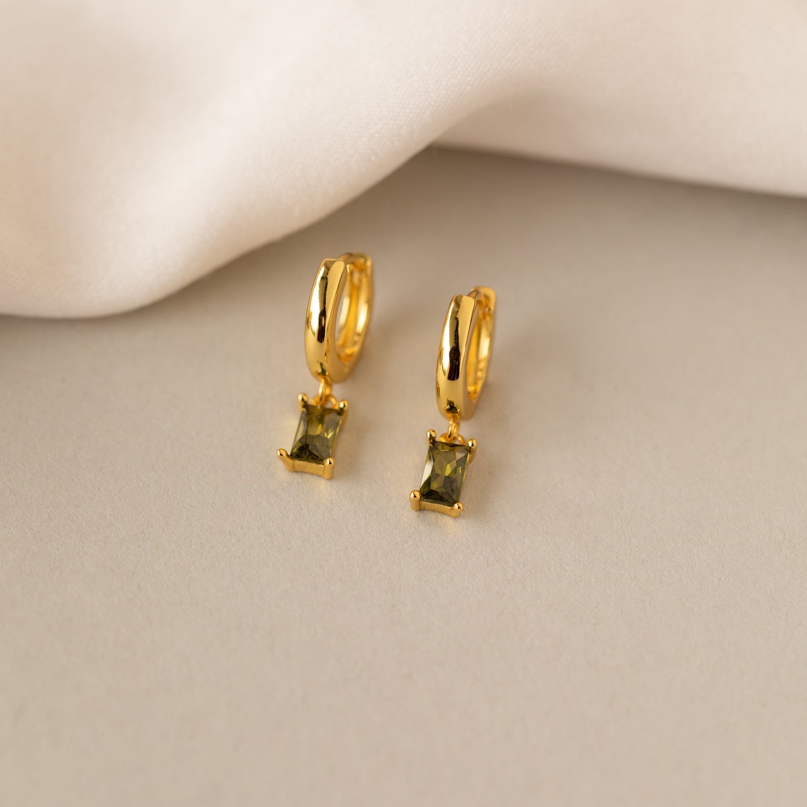 Baguette Birthstone Huggies—gold hoop earrings with rectangular green gemstones—are displayed on a light beige surface near soft white fabric, perfect for adding personalized style.