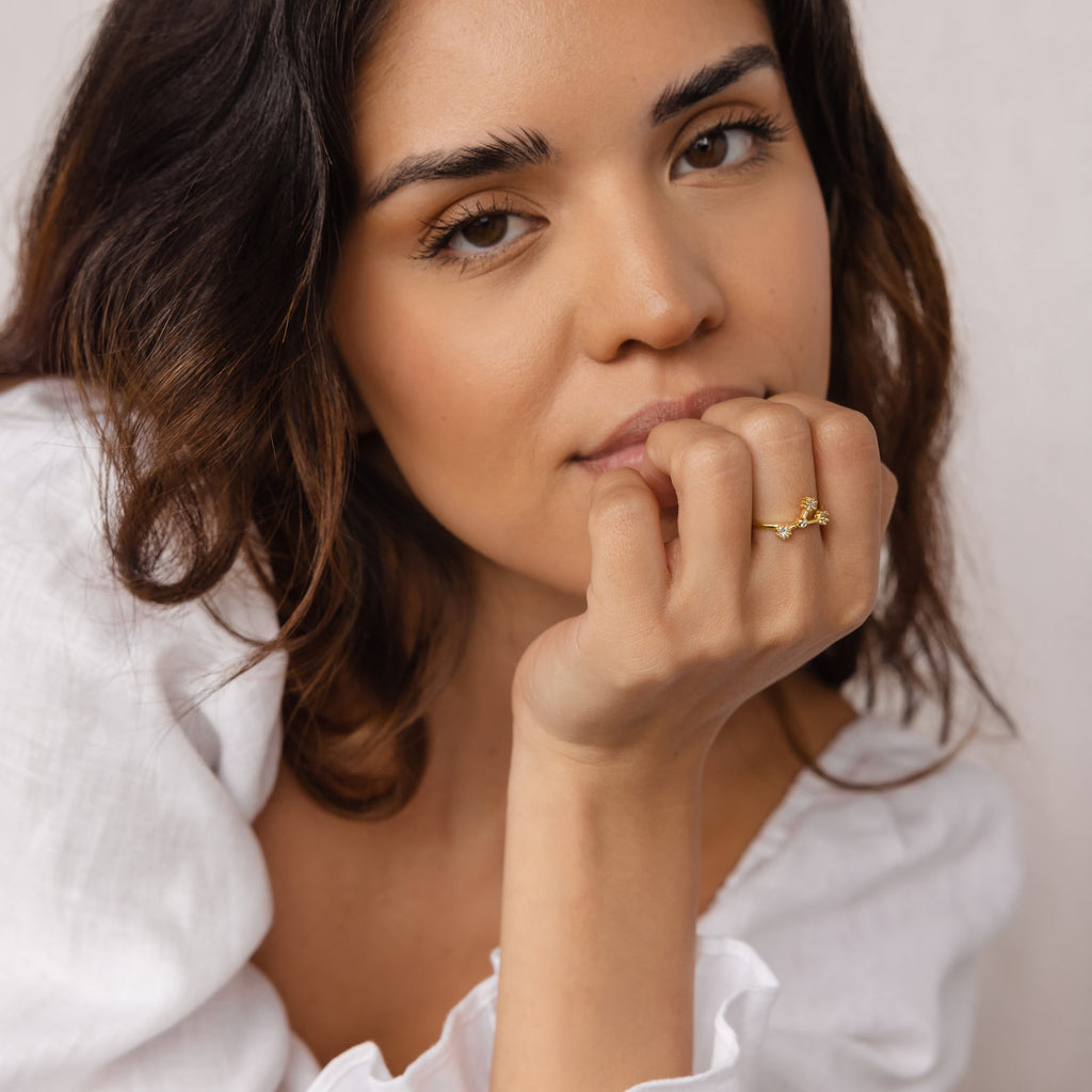 A woman with wavy hair in a white top rests her chin on her hand, wearing the Constellation Ring for a subtle celestial touch that enhances her elegant style.
