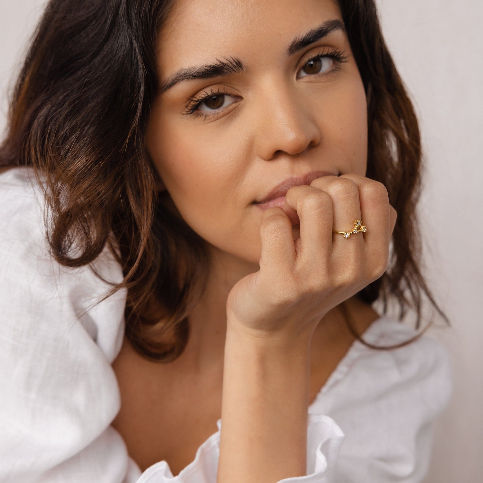 A woman with wavy hair in a white top rests her chin on her hand, wearing the Constellation Ring for a subtle celestial touch that enhances her elegant style.