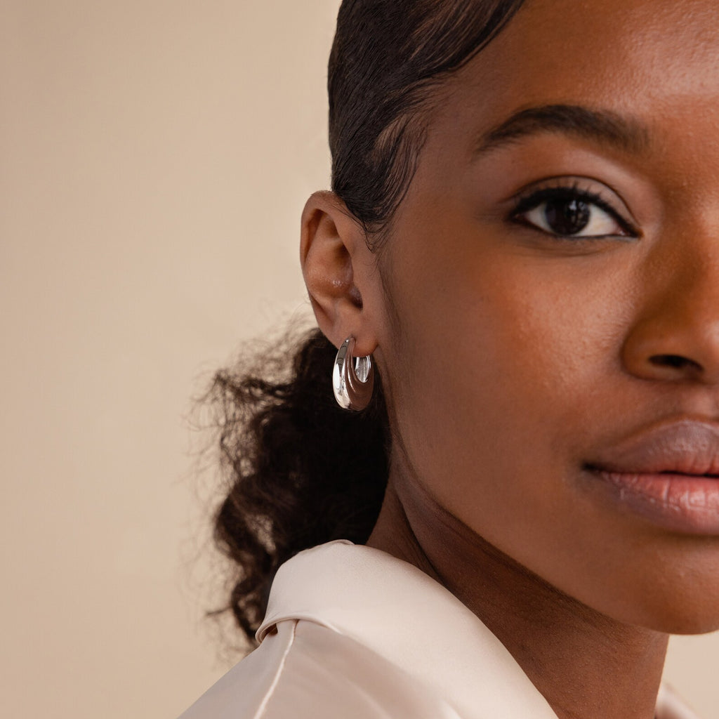 Close-up of a woman with curly hair wearing a cream blouse and Katrina Ribbed Hoops featuring a chunky ribbed texture and modern design, looking at the camera.
