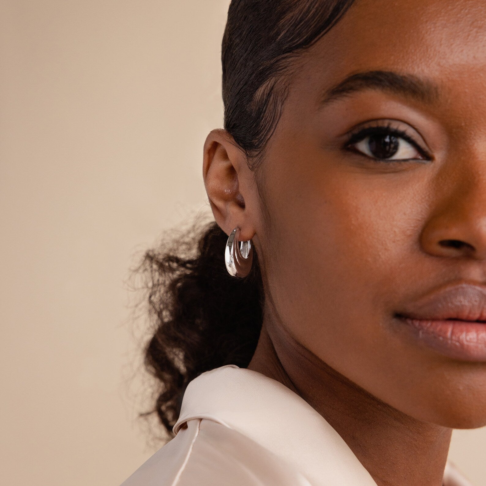 Close-up of a woman with curly hair wearing a cream blouse and Katrina Ribbed Hoops featuring a chunky ribbed texture and modern design, looking at the camera.
