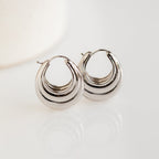 The Katrina Ribbed Hoops are shiny silver statement oval earrings with a chunky, layered ribbed design, displayed on a white surface.