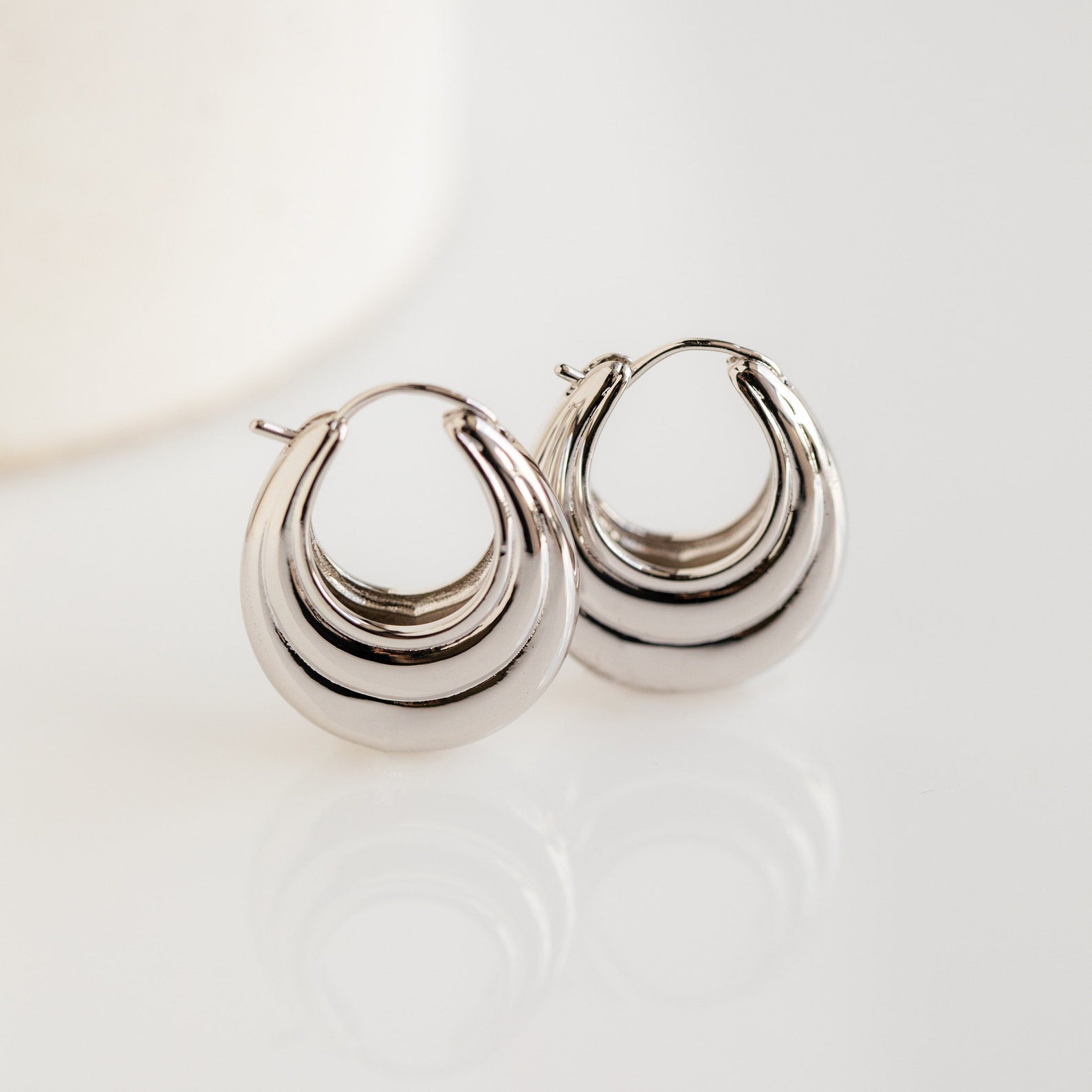 The Katrina Ribbed Hoops are shiny silver statement oval earrings with a chunky, layered ribbed design, displayed on a white surface.