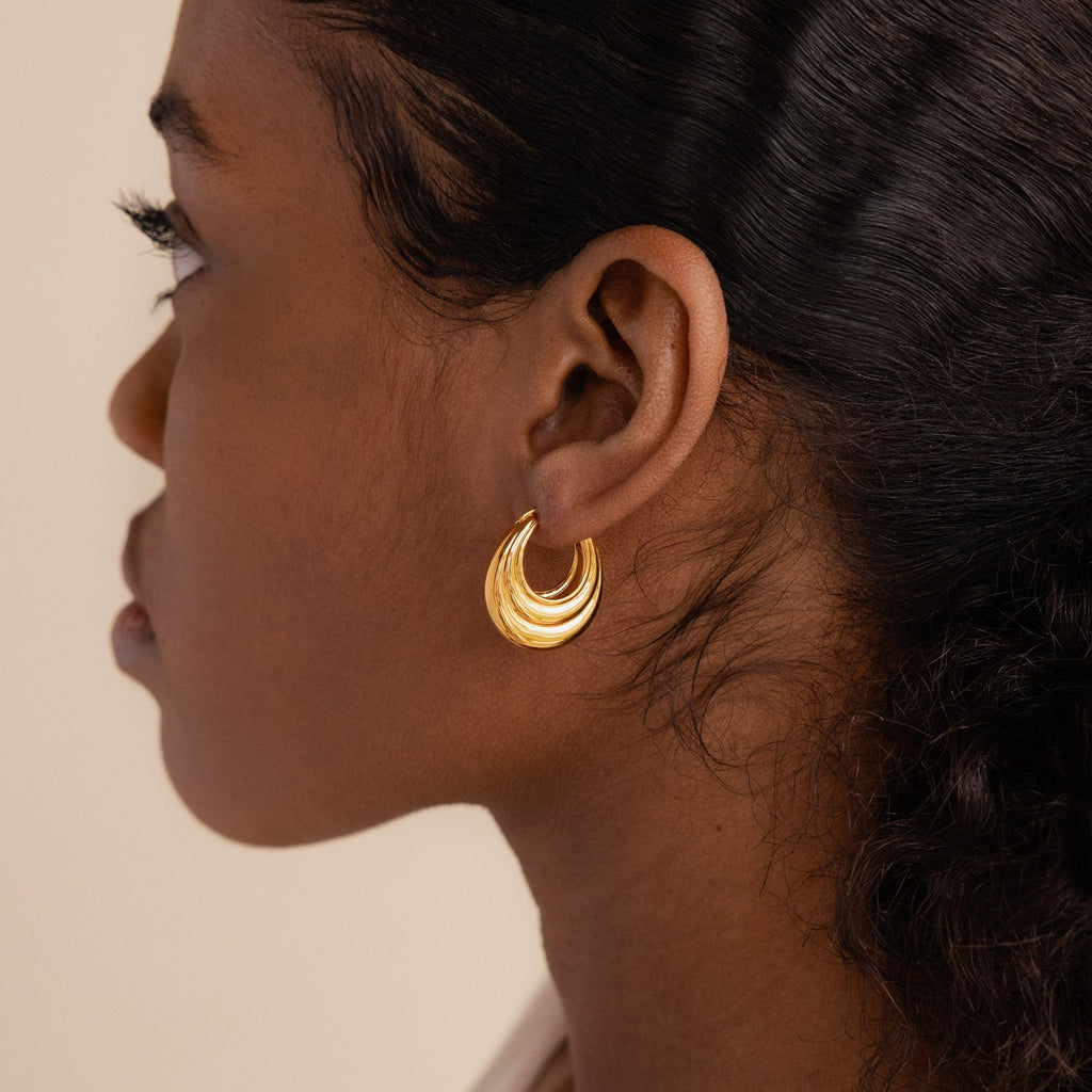 A woman with dark hair wears Katrina Ribbed Hoops, chunky oval earrings with a ribbed texture, shown in profile against a neutral background.