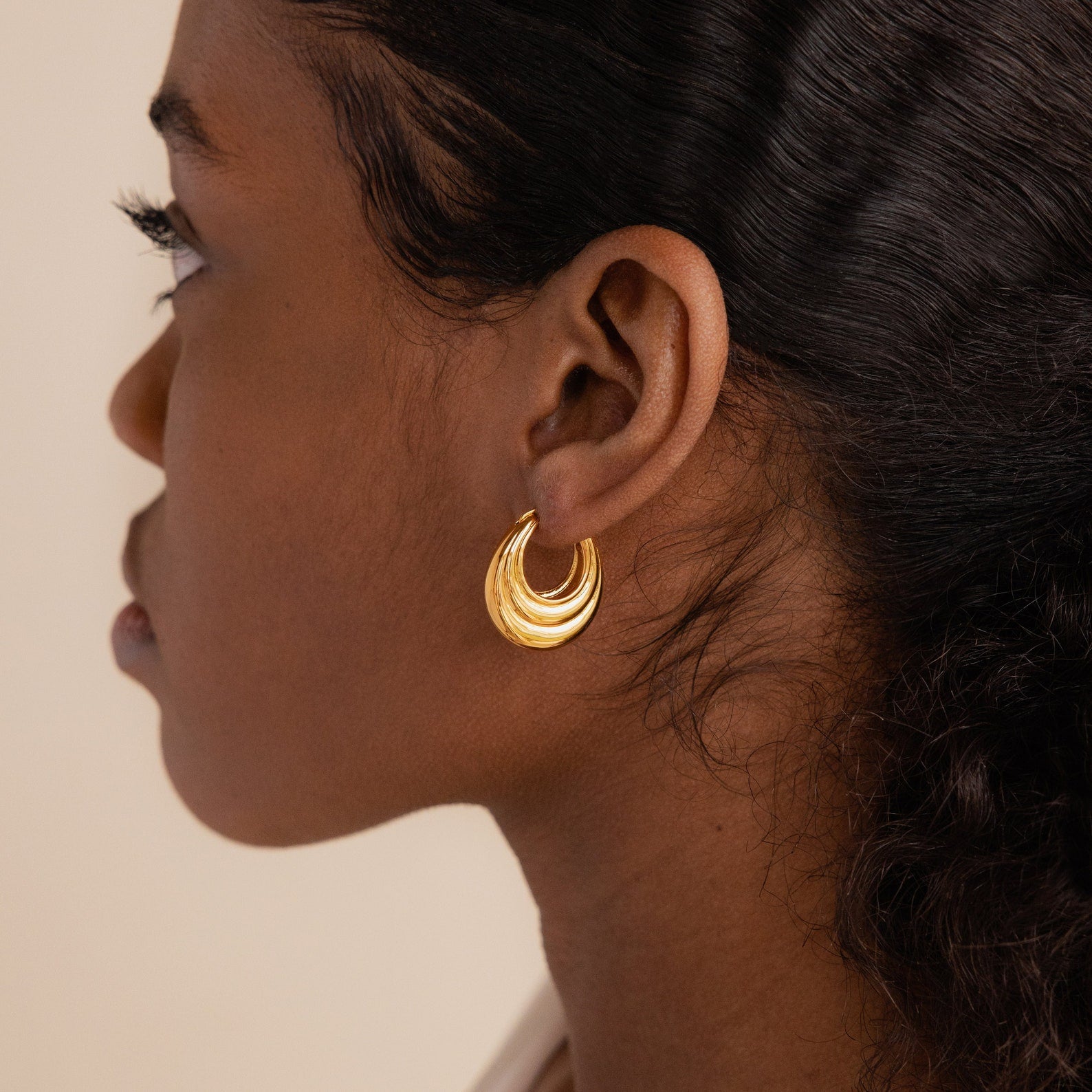 A woman with dark hair wears Katrina Ribbed Hoops, chunky oval earrings with a ribbed texture, shown in profile against a neutral background.