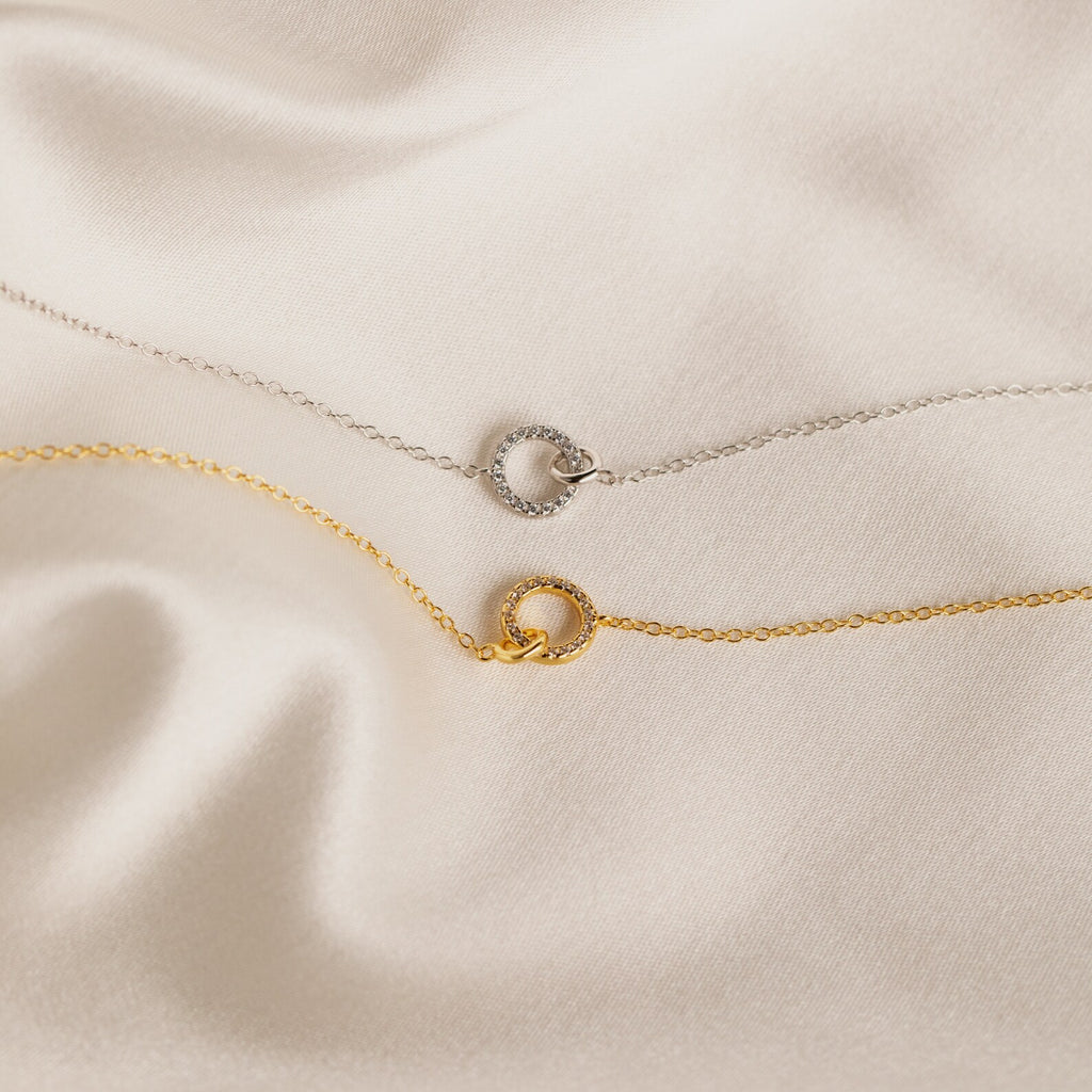 A gold and a silver Pave Interlocking Necklace are displayed on cream-colored satin fabric—perfect as a wedding gift or for lovers of minimal diamond jewelry.