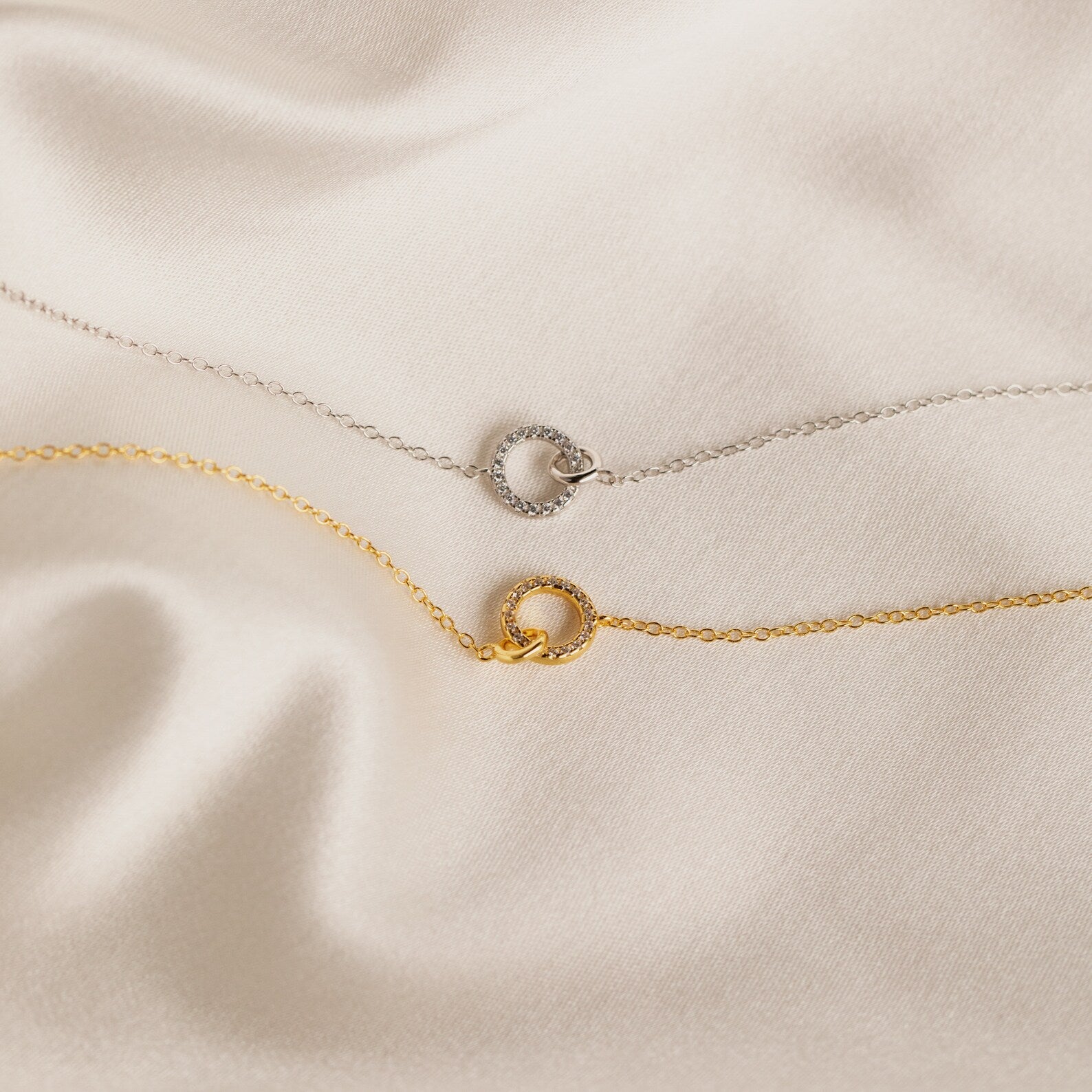 A gold and a silver Pave Interlocking Necklace are displayed on cream-colored satin fabric—perfect as a wedding gift or for lovers of minimal diamond jewelry.
