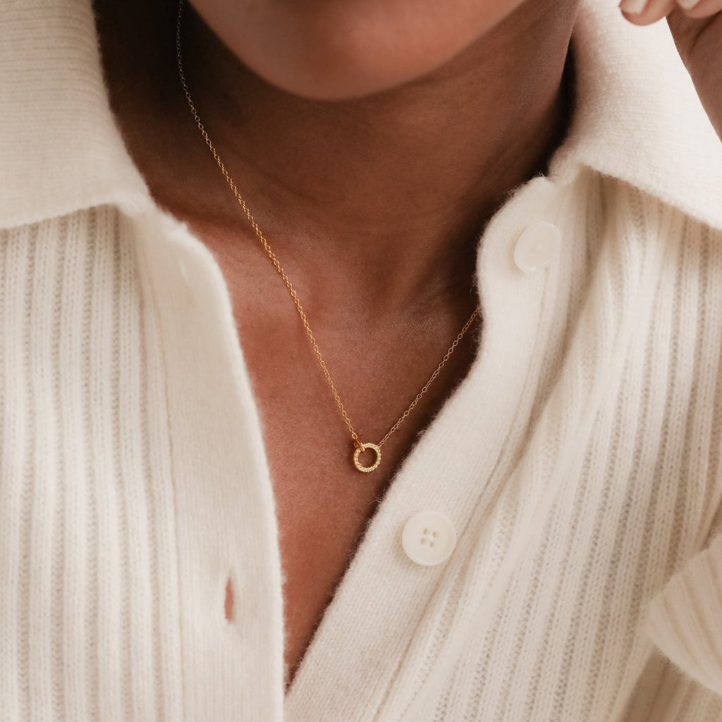 A person wearing the Pave Interlocking Necklace with a gold circle pendant and a cream, ribbed button-up shirt—an elegant style ideal as a wedding gift.