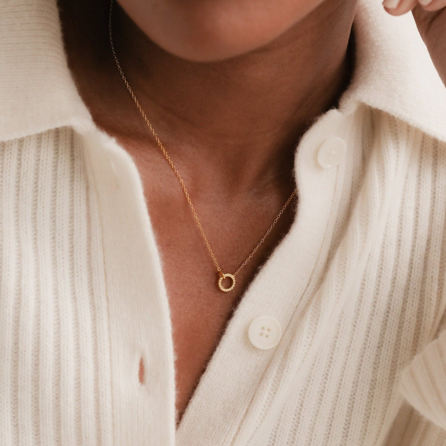 A person wearing the Pave Interlocking Necklace with a gold circle pendant and a cream, ribbed button-up shirt—an elegant style ideal as a wedding gift.