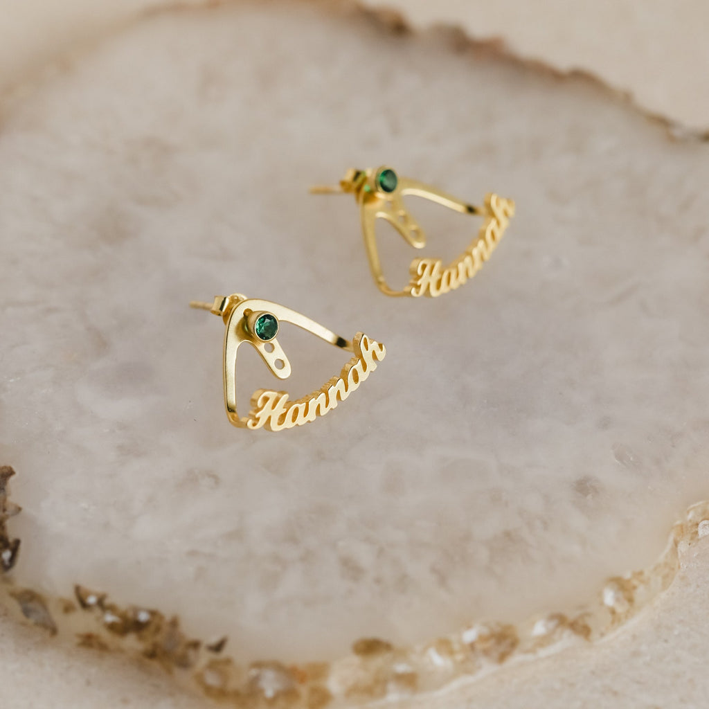 Featuring green birthstones and a personalized Hannah nameplate, the London Name Ear Jackets are stylish wrap-around earrings shown on a textured beige surface.