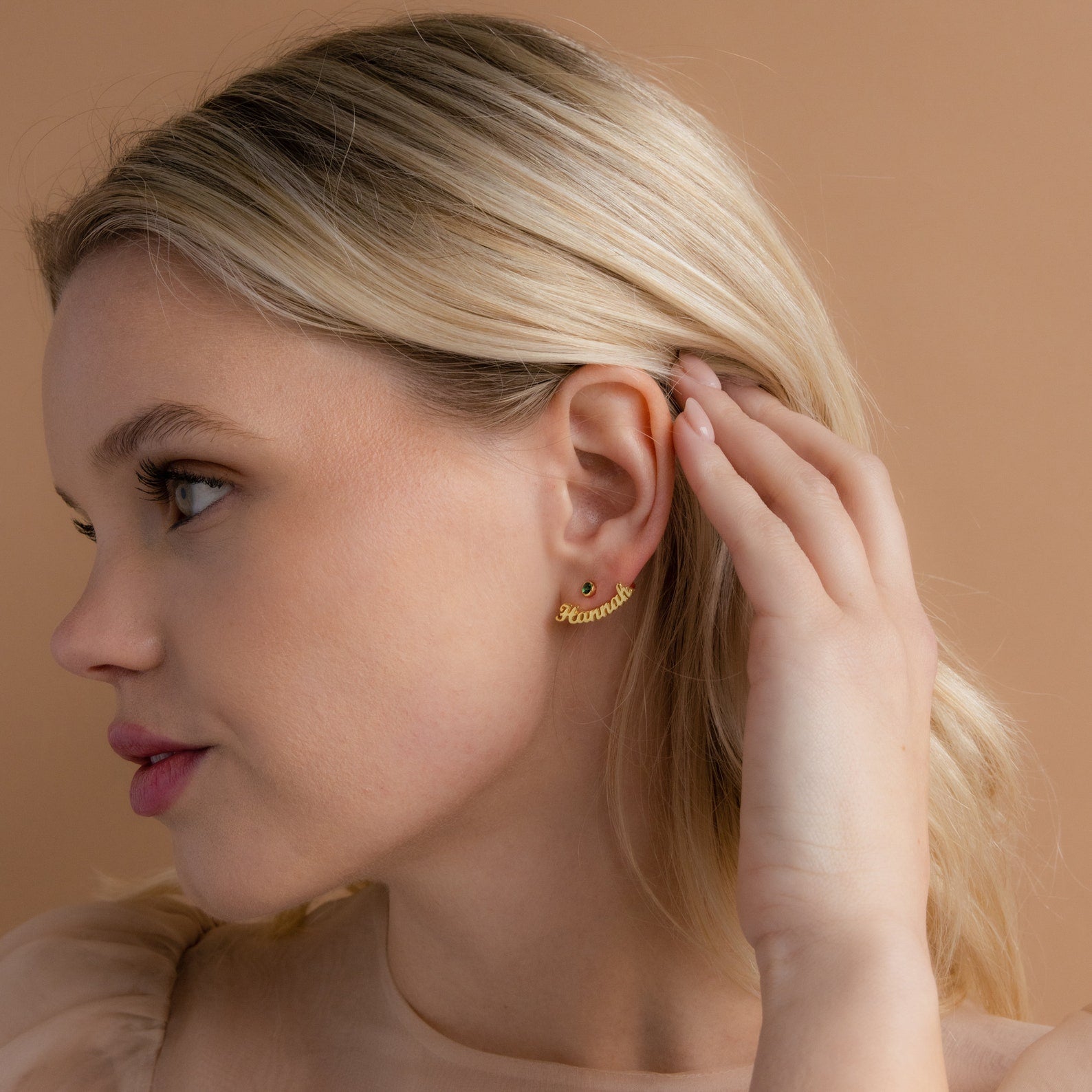 A blonde woman touches her ear, wearing London Name Ear Jackets personalized with "Rihanna," set against a beige background.