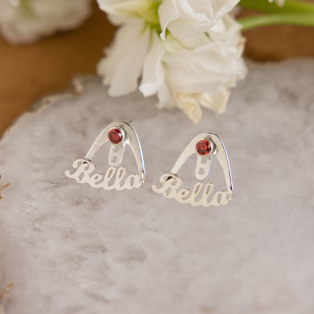 The London Name Ear Jackets, custom birthstone earrings spelling "Bella" with a red gemstone, are shown on a stone slab beside white flowers.