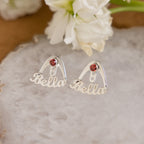 The London Name Ear Jackets, custom birthstone earrings spelling "Bella" with a red gemstone, are shown on a stone slab beside white flowers.