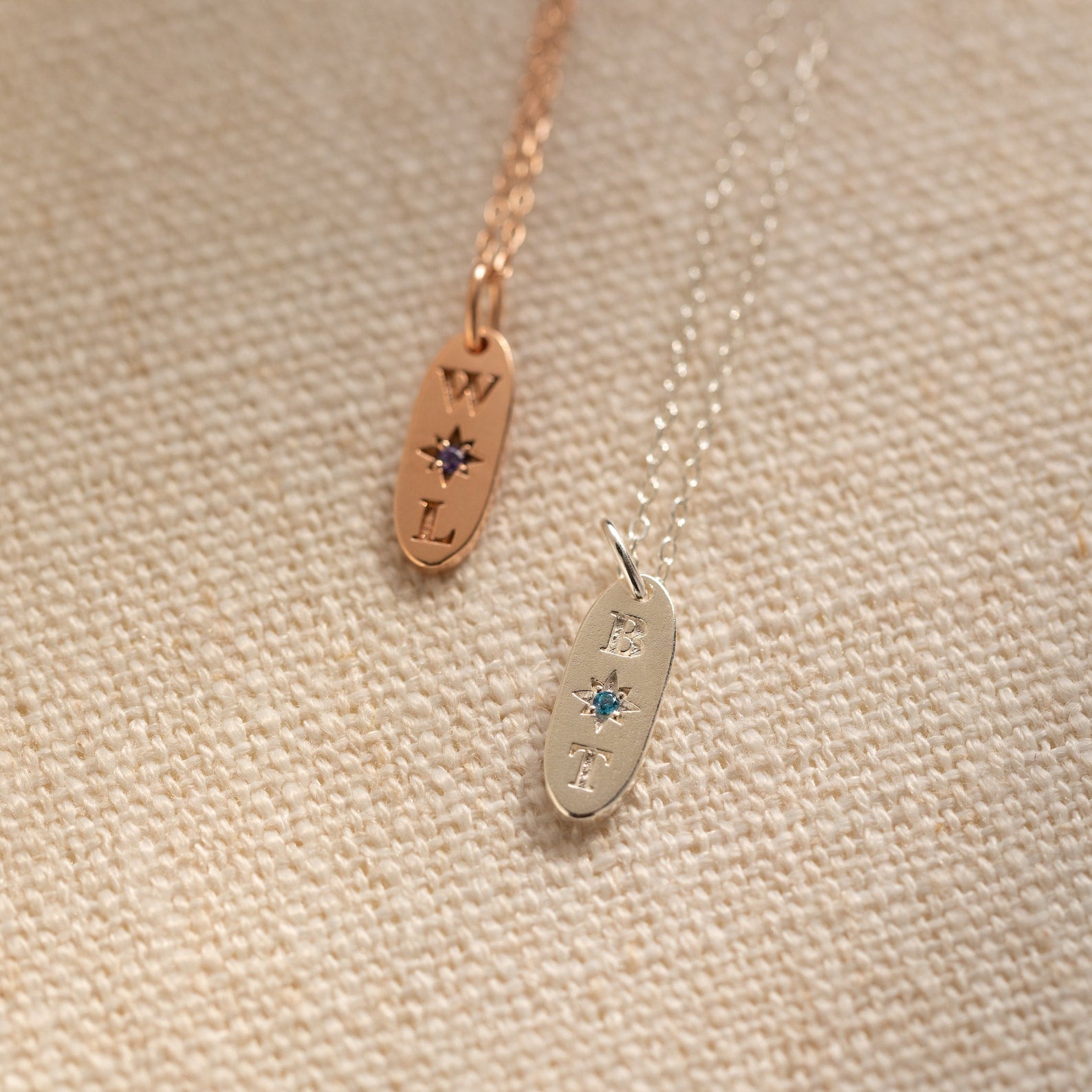 Two Newsletter Oval Tag Necklaces, each featuring an engraved letter and a star-shaped birthstone, rest on a textured surface.