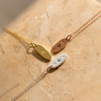 The Newsletter Oval Tag Necklace is shown in gold, rose gold, and silver on a beige stone surface—each personalized necklace adds timeless elegance and meaning.