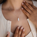 A woman wears the Newsletter Oval Tag Necklace, a silver personalized piece featuring an engraved pendant, as she touches her chest with manicured hands.