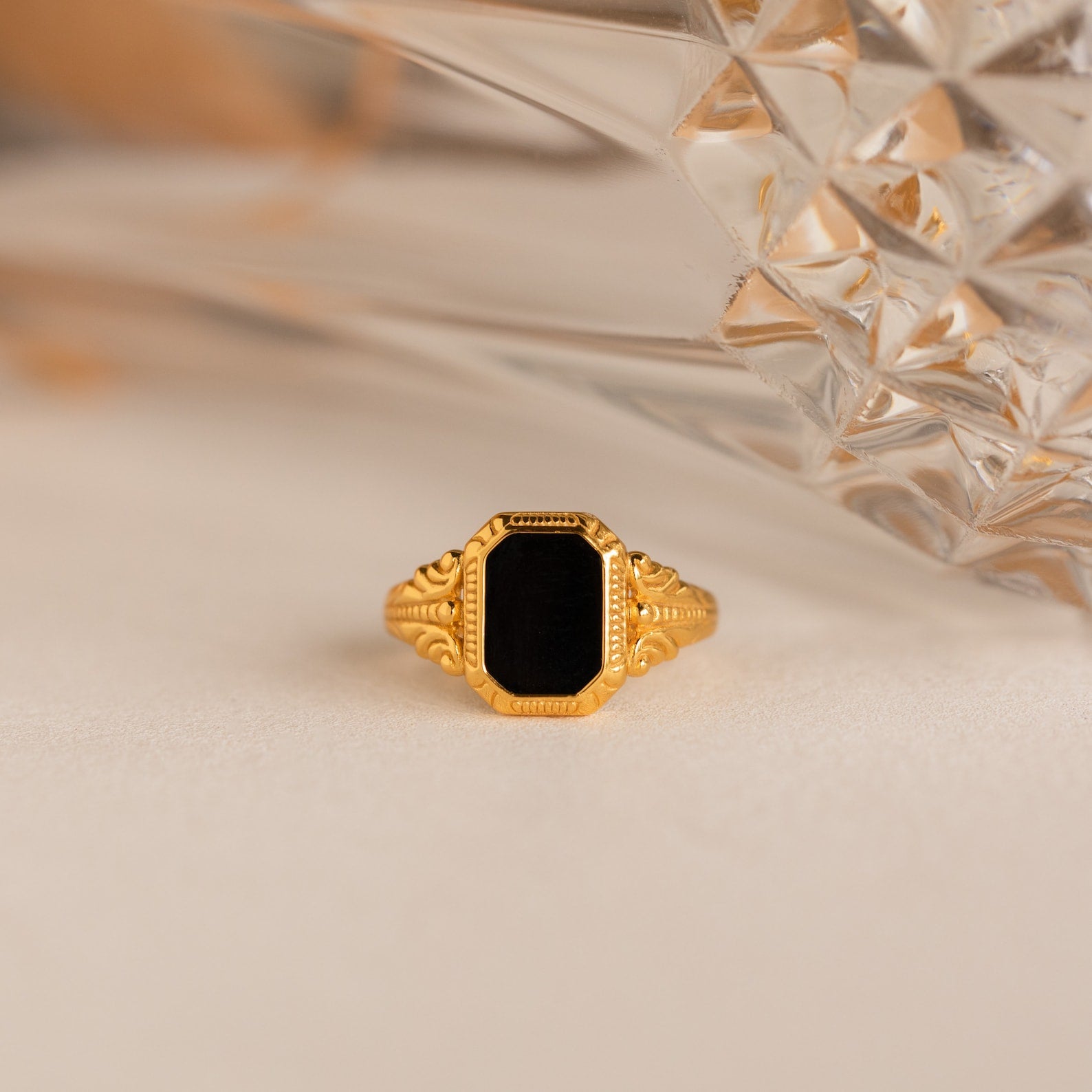 The Vintage Black Signet Ring features an ornate gold band and a rectangular black stone, displayed on a light surface beside crystal decor.