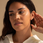 A woman with long dark hair touches her ear, elegantly displaying the Halo Opal Hoops while wearing a white top and gazing thoughtfully to the side.