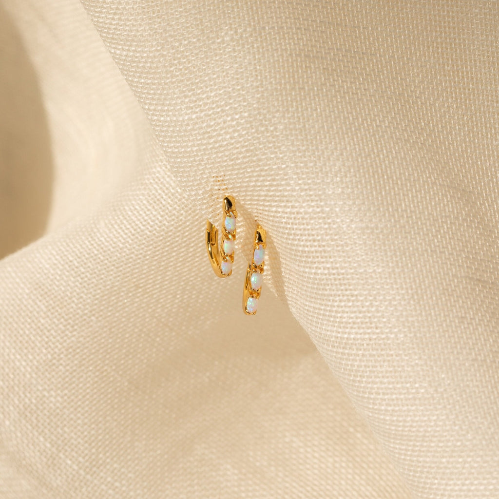 Halo Opal Hoops—gold huggie earrings featuring petite white opals, shown on textured cream fabric for a chic, minimalist style.