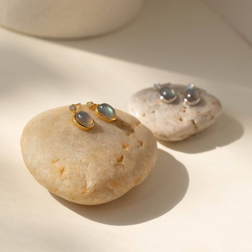 Two pairs of Moonstone Drop Earrings—one gold, one silver—rest on smooth stones in soft natural light. These minimalist earrings are an elegant graduation gift or a timeless addition to your collection.