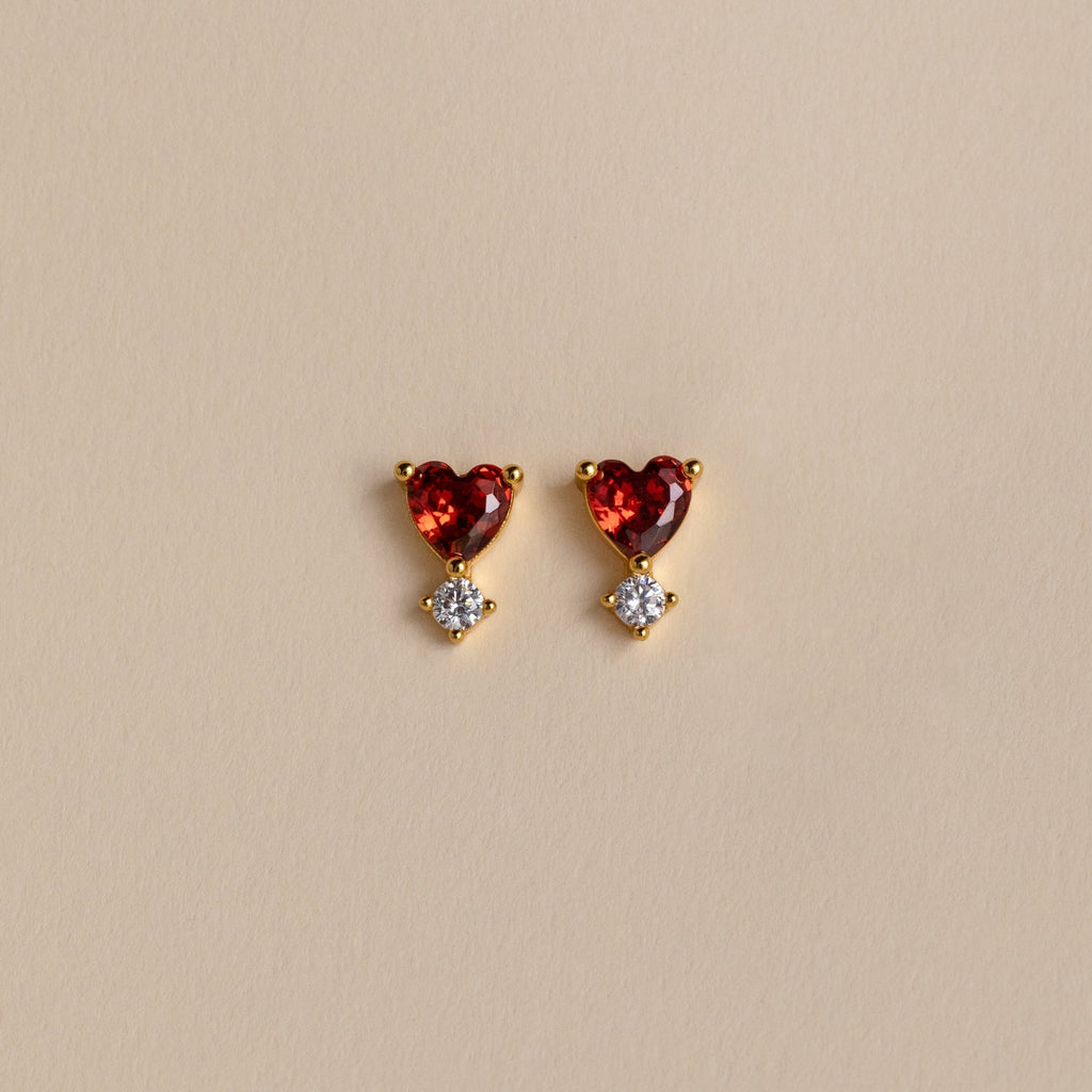 Garnet Heart Studs feature gold settings with heart-shaped garnet gems above small round clear stones, displayed on a beige background—delicate earrings that bring a romantic touch to any look.