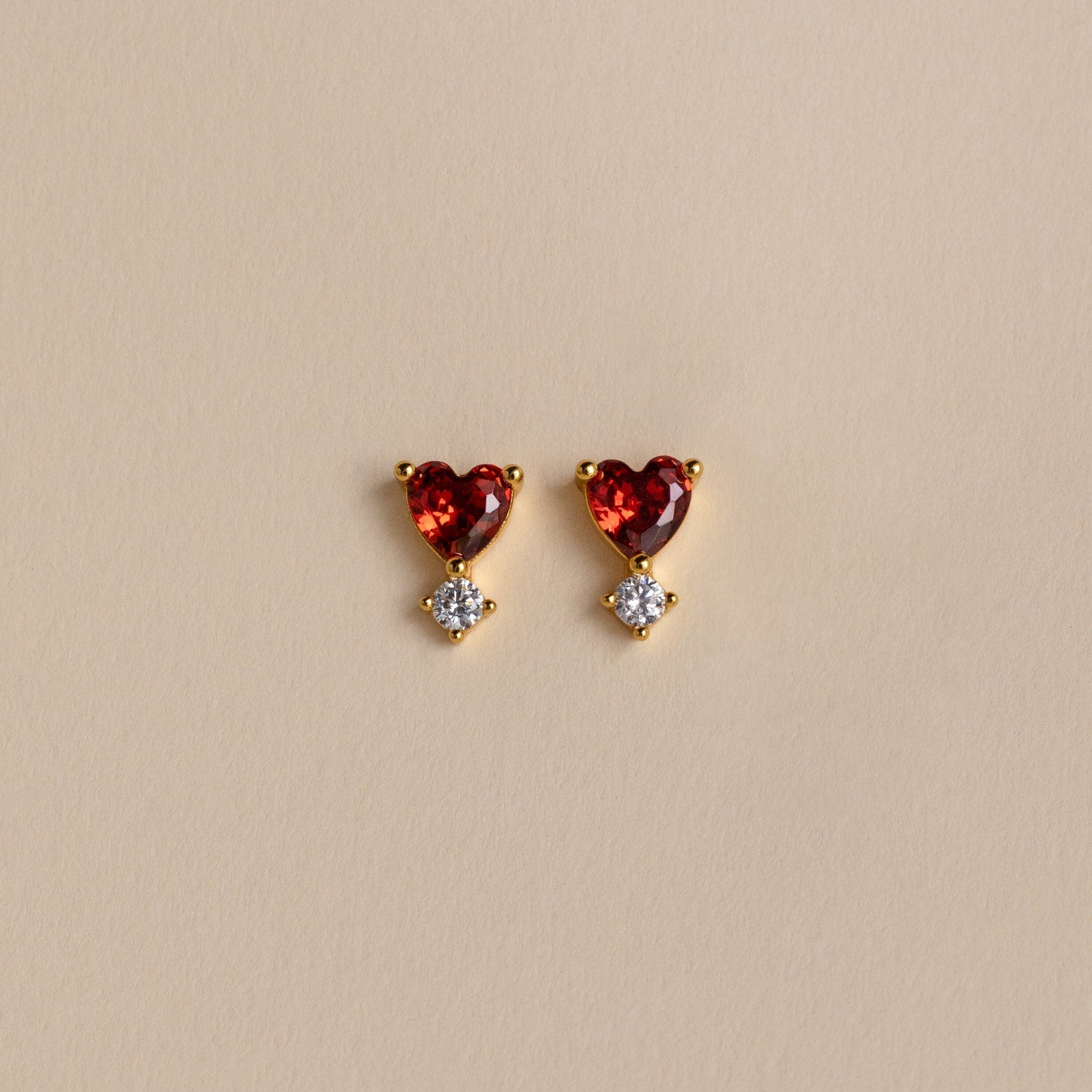 Garnet Heart Studs feature gold settings with heart-shaped garnet gems above small round clear stones, displayed on a beige background—delicate earrings that bring a romantic touch to any look.