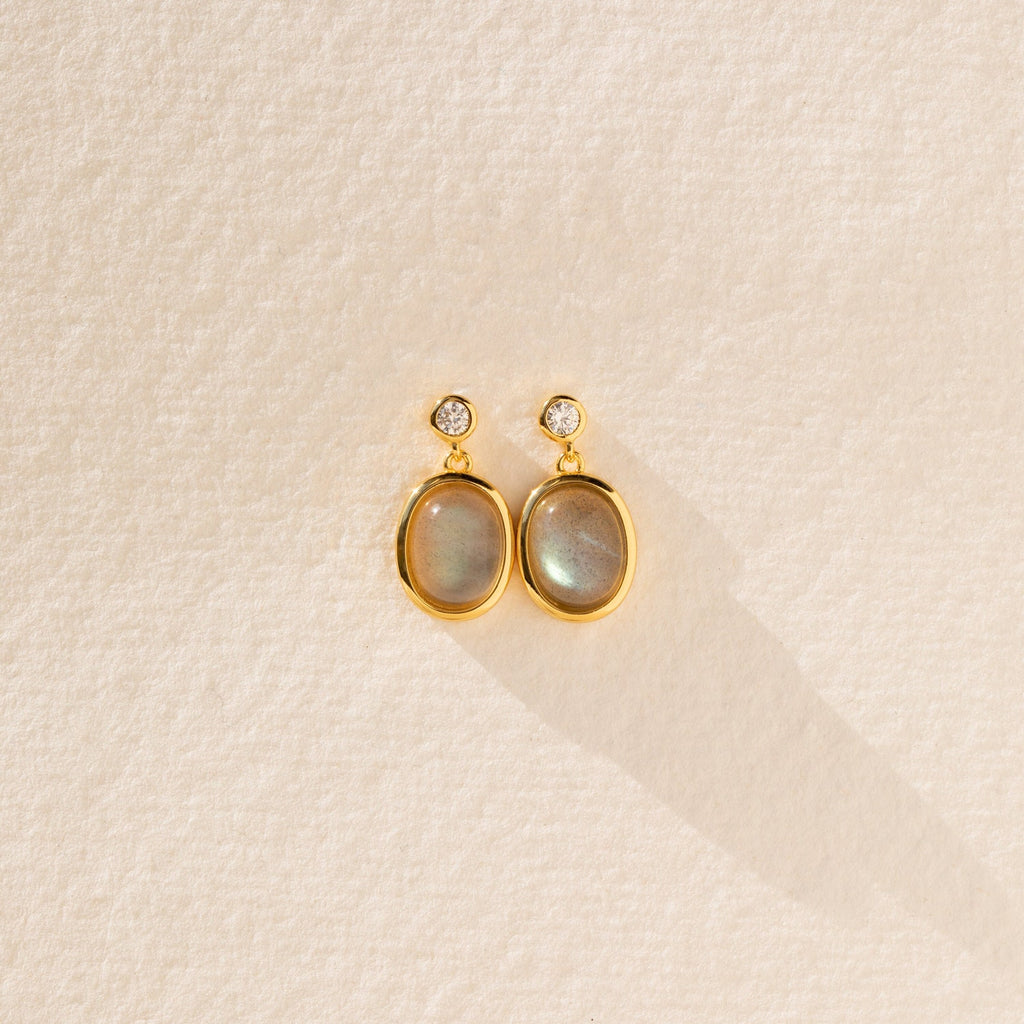 Moonstone Drop Earrings feature gold-framed oval moonstones with small round accents, displayed on a textured beige surface—a perfect minimalist accessory and thoughtful graduation gift.