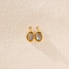 Moonstone Drop Earrings feature gold-framed oval moonstones with small round accents, displayed on a textured beige surface—a perfect minimalist accessory and thoughtful graduation gift.