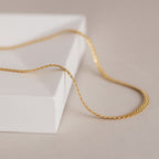 The Men's Snake Chain Necklace, a delicate gold chain ideal for minimalist style or layering with men's jewelry, is displayed on a white textured surface.